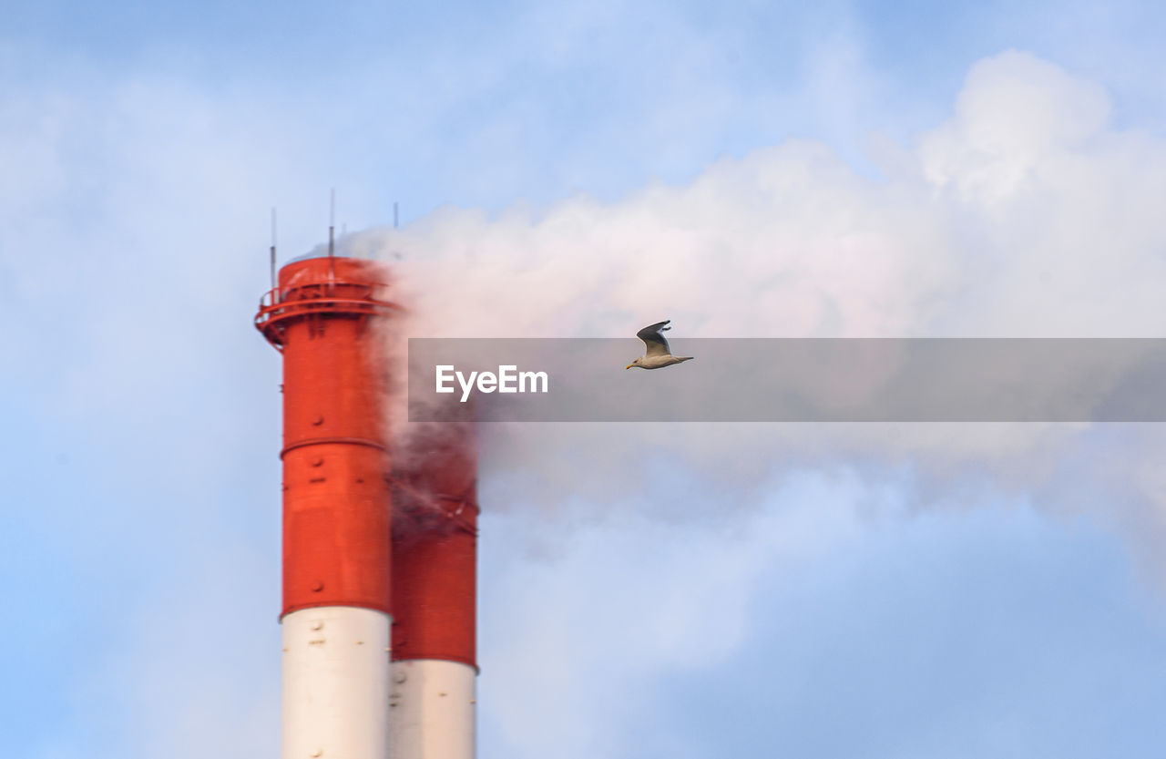 sky, smoke, flying, cloud, tower, nature, smoke stack, architecture, day, no people, building exterior, industry, built structure, factory, low angle view, outdoors, lighthouse, pollution, red, environmental issues, transportation, motion, blue, air vehicle, bird, air pollution, fumes, airplane