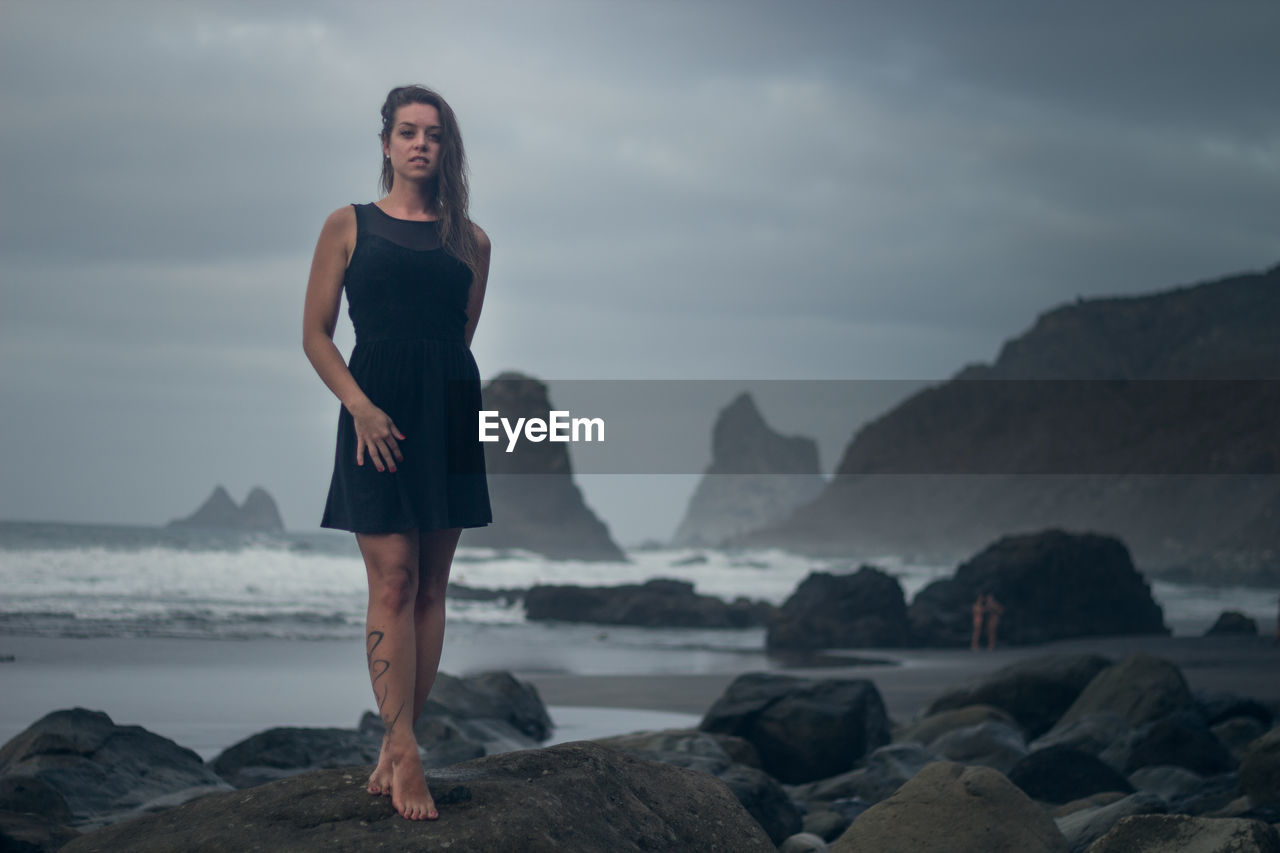 Portrait of young woman standing on rock by sea against sky