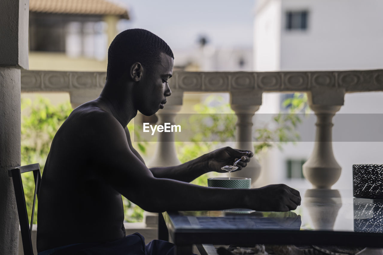 Seated shirtless on a hotel terrace, an african american man savors breakfast food with a side view