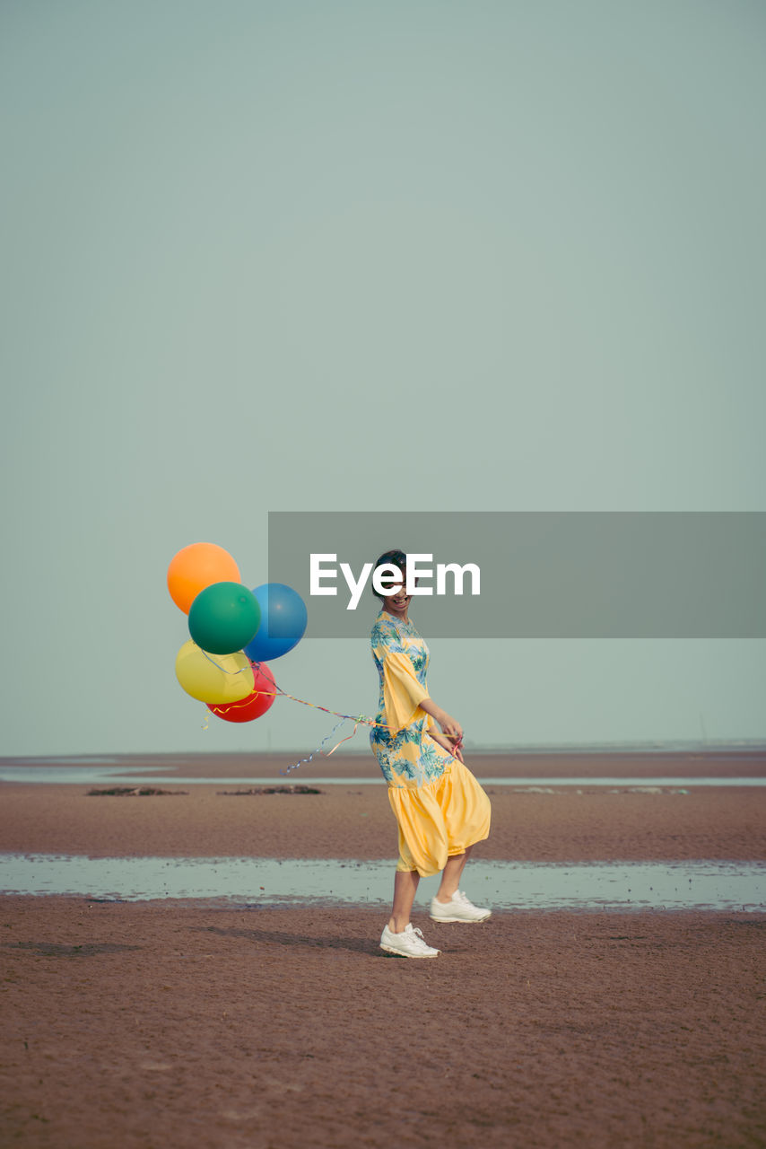 Full length of young woman holding colorful balloons at beach