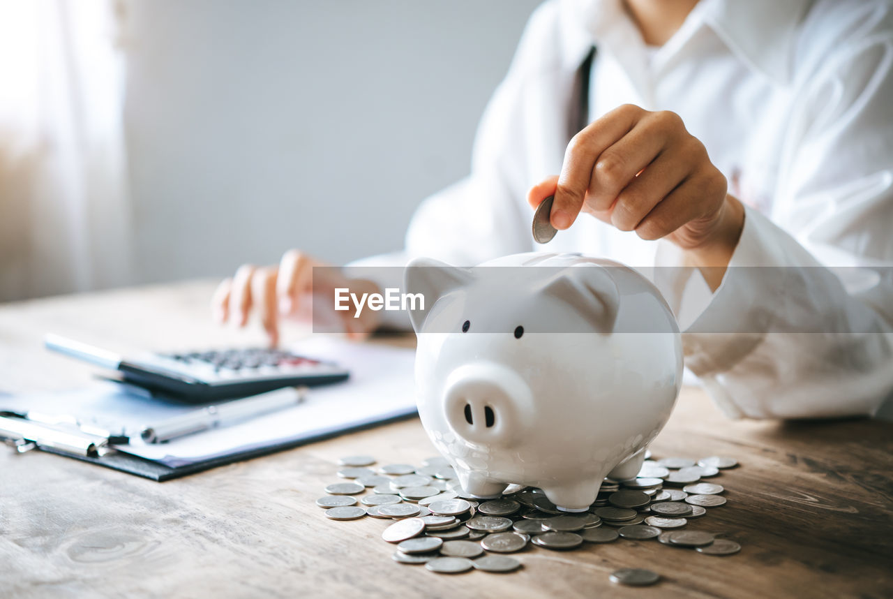 midsection of businesswoman putting coin in piggy bank on table