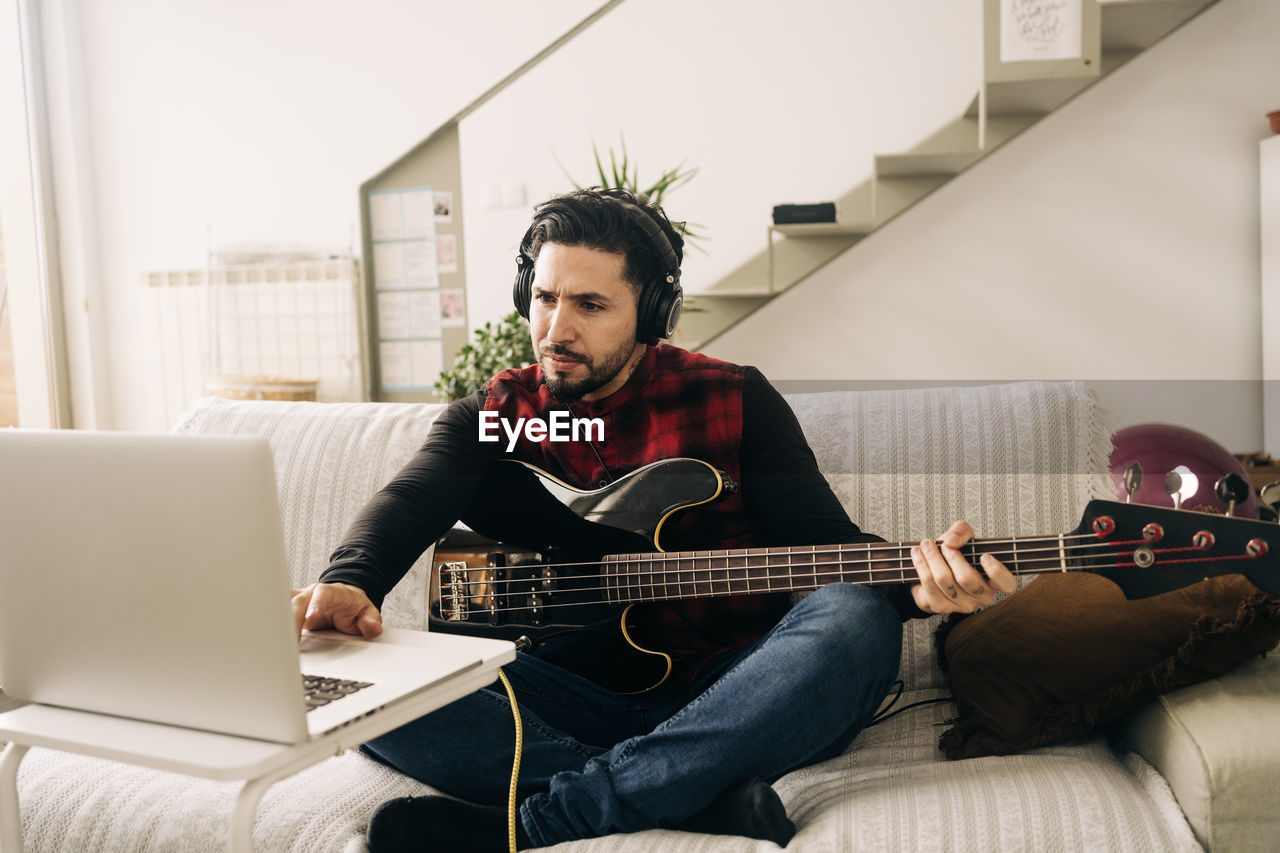 Adult male musician in headphones playing bass guitar against netbook on sofa in living room