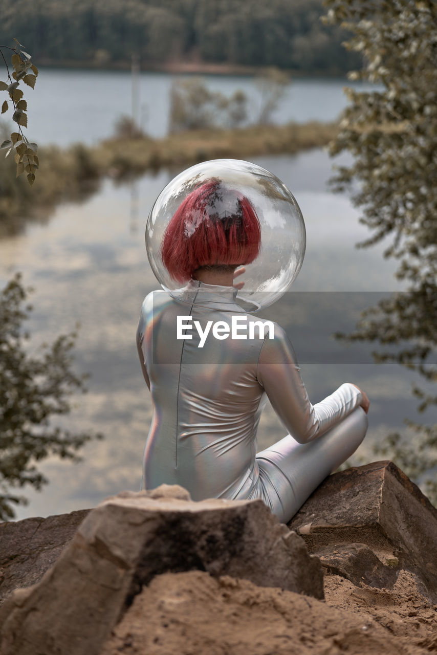 Back view of unrecognizable futuristic young red haired female in silver space suit and glass helmet sitting near flooding river