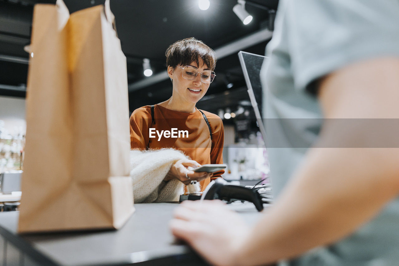 Smiling mature woman making payment through smart phone at checkout counter in electronics store