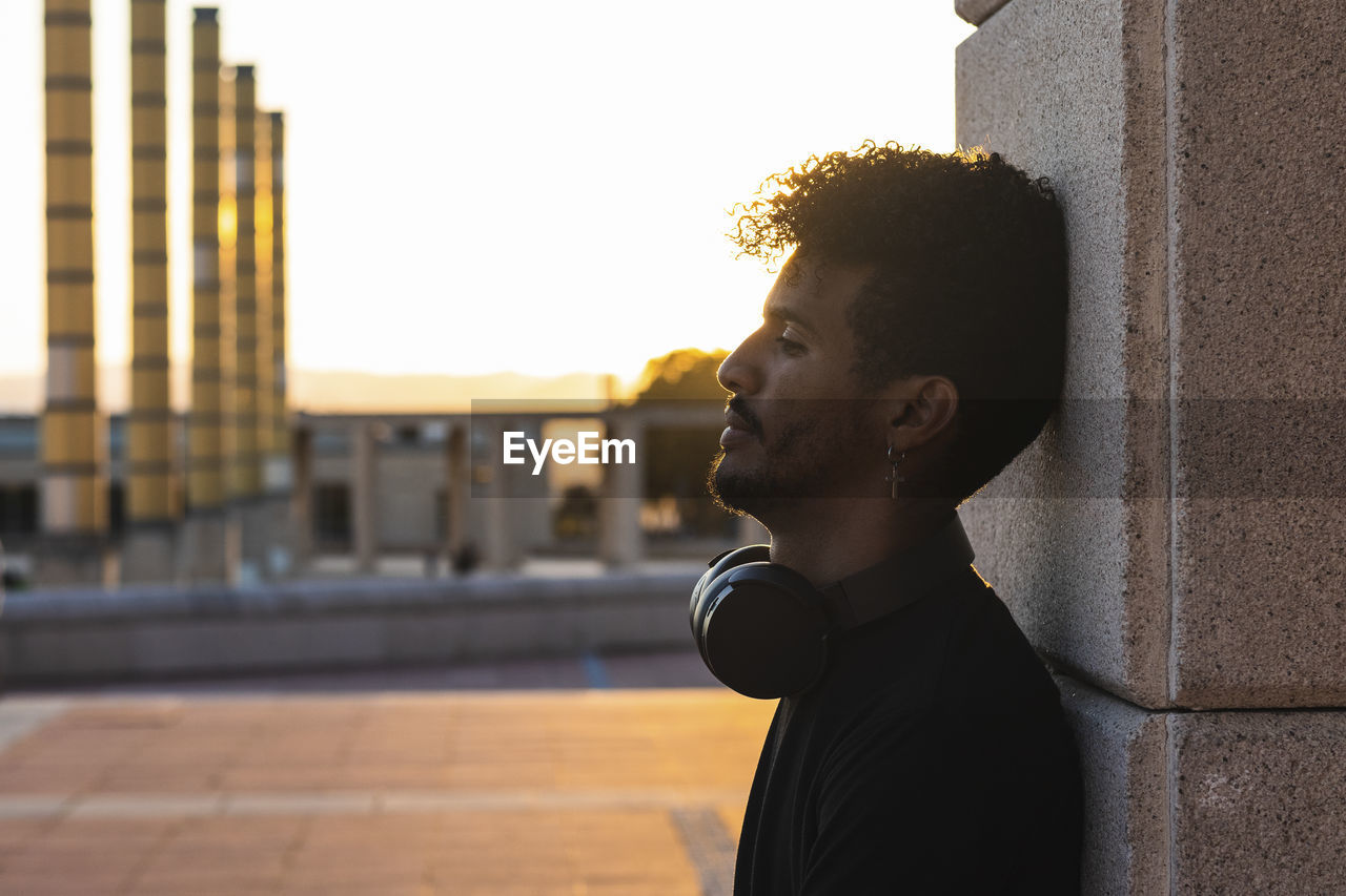Thoughtful young man with headphones leaning on wall against clear sky during sunset