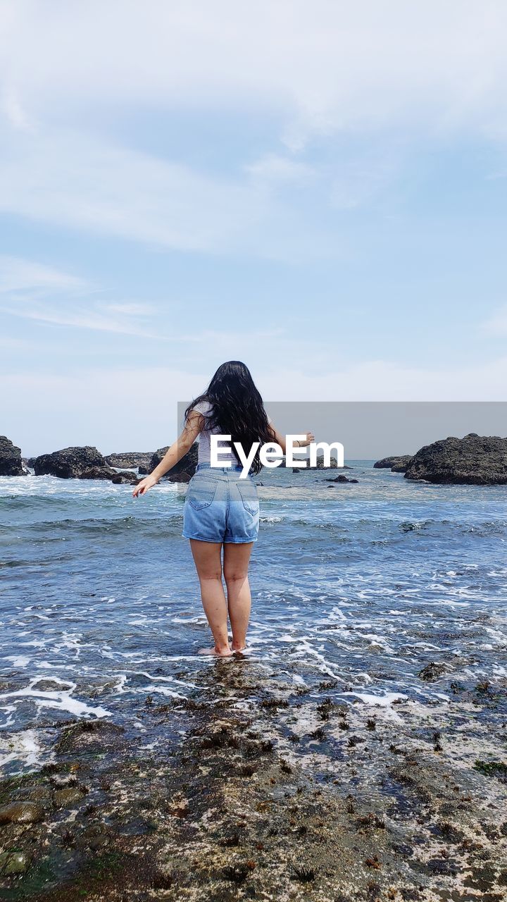 REAR VIEW OF WOMAN STANDING ON BEACH AGAINST SEA