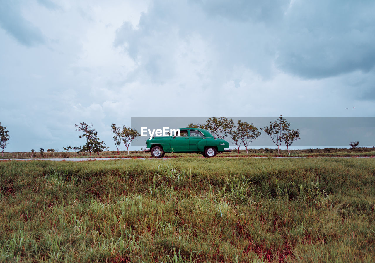 Sandy dirt red road with old vintage car with green plants on sides and grey cloudy sky on background in cuba