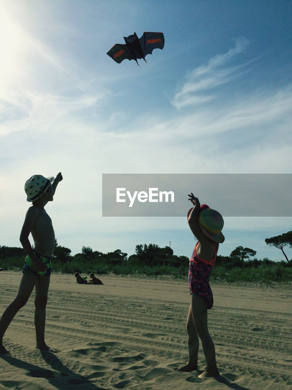 Full length of boy with sister flying kite at sandy beach against sky