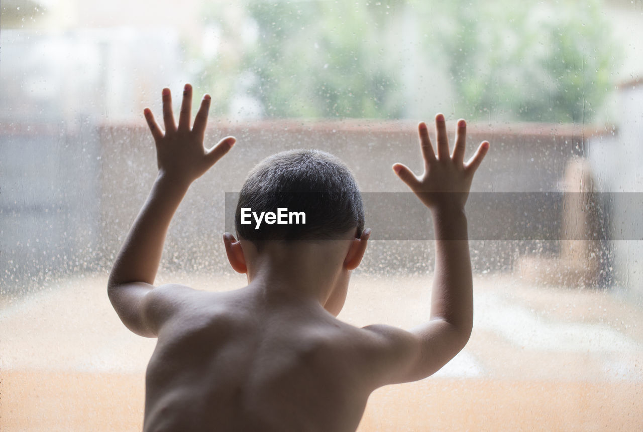Rear view of shirtless boy looking through wet glass window