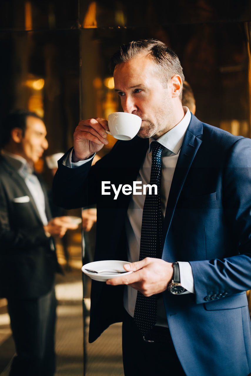 Mature businessman drinking coffee while standing against reflection of male colleague on glass