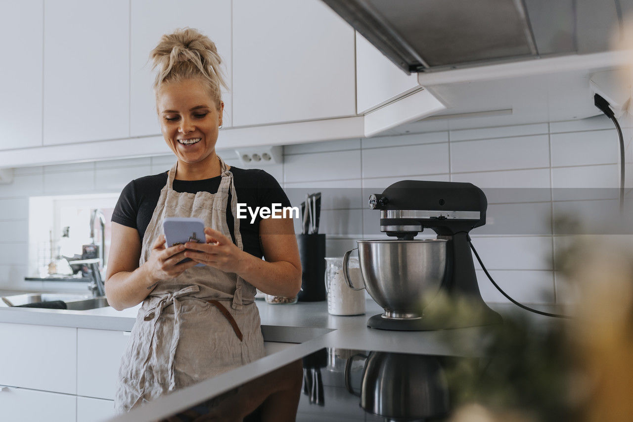 Smiling young woman in kitchen using cell phone