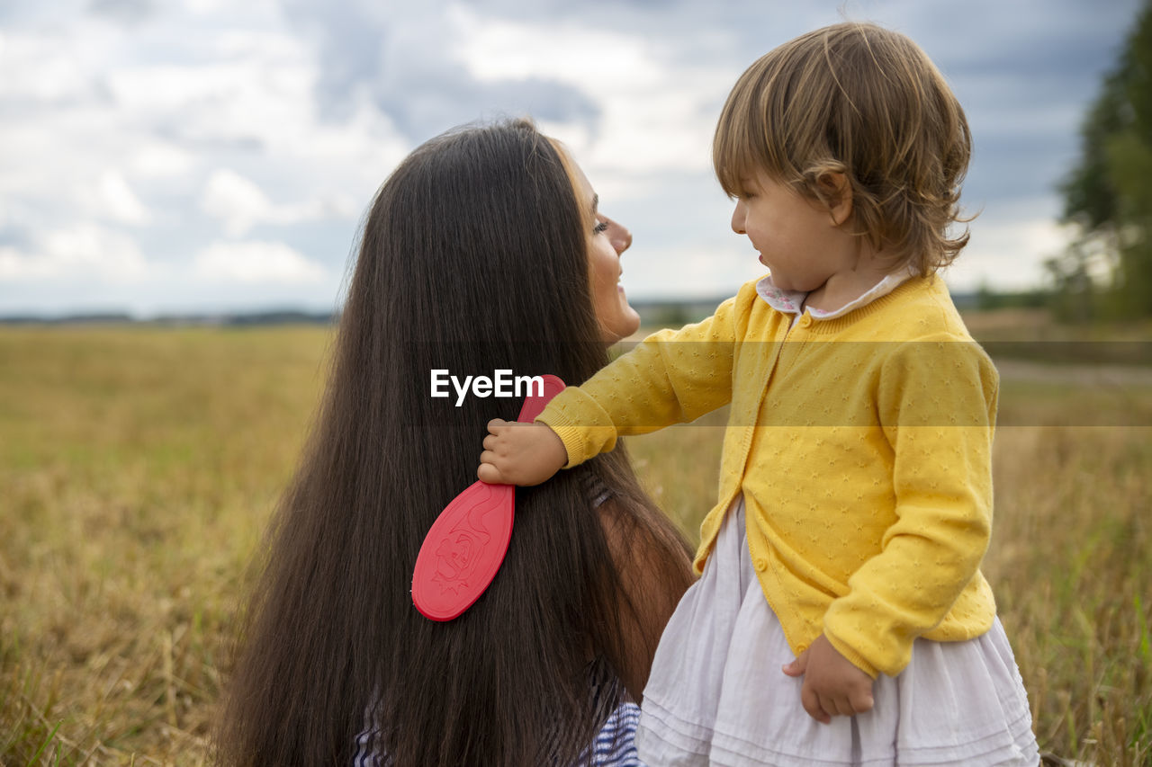 Adorable toddler girl play combing her mom's hair outdoor. rear view of mother and daughter on field