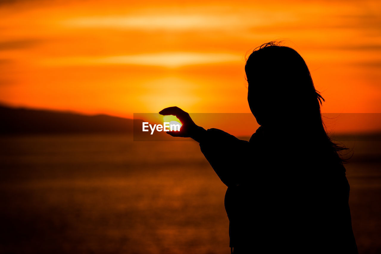 Optical illusion of silhouette woman holding sun at beach during sunset