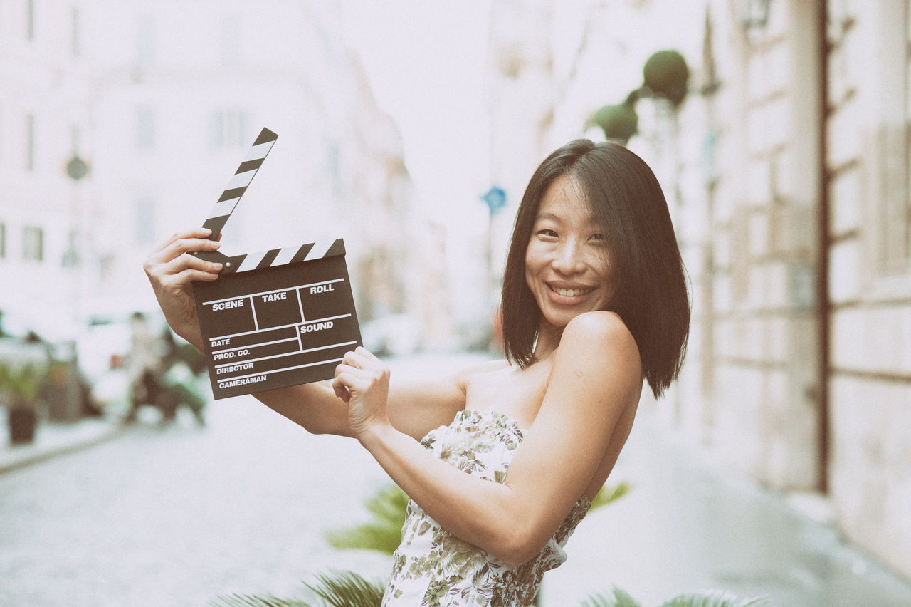 Portrait of smiling woman holding film slate while standing in city