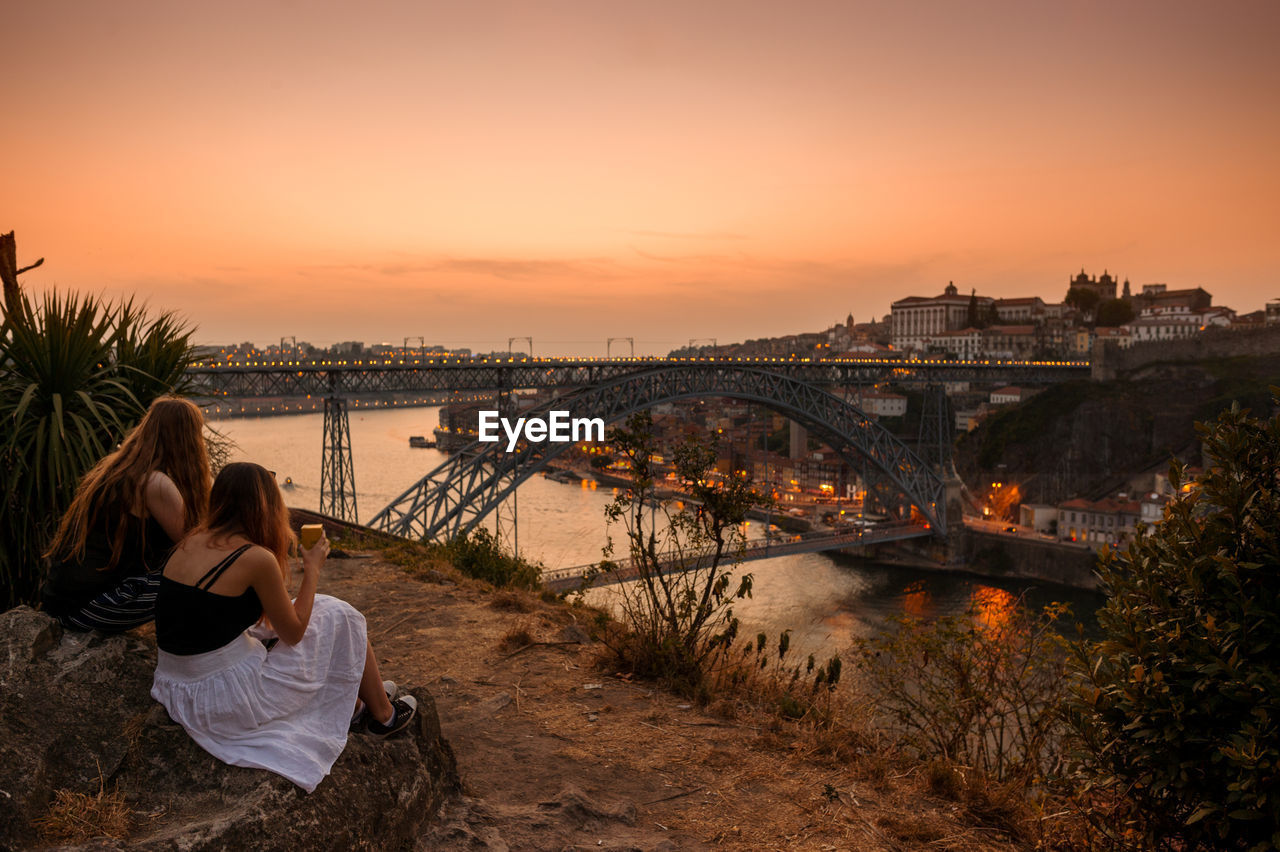 Rear view of women looking at bridge over river against orange sky during sunset