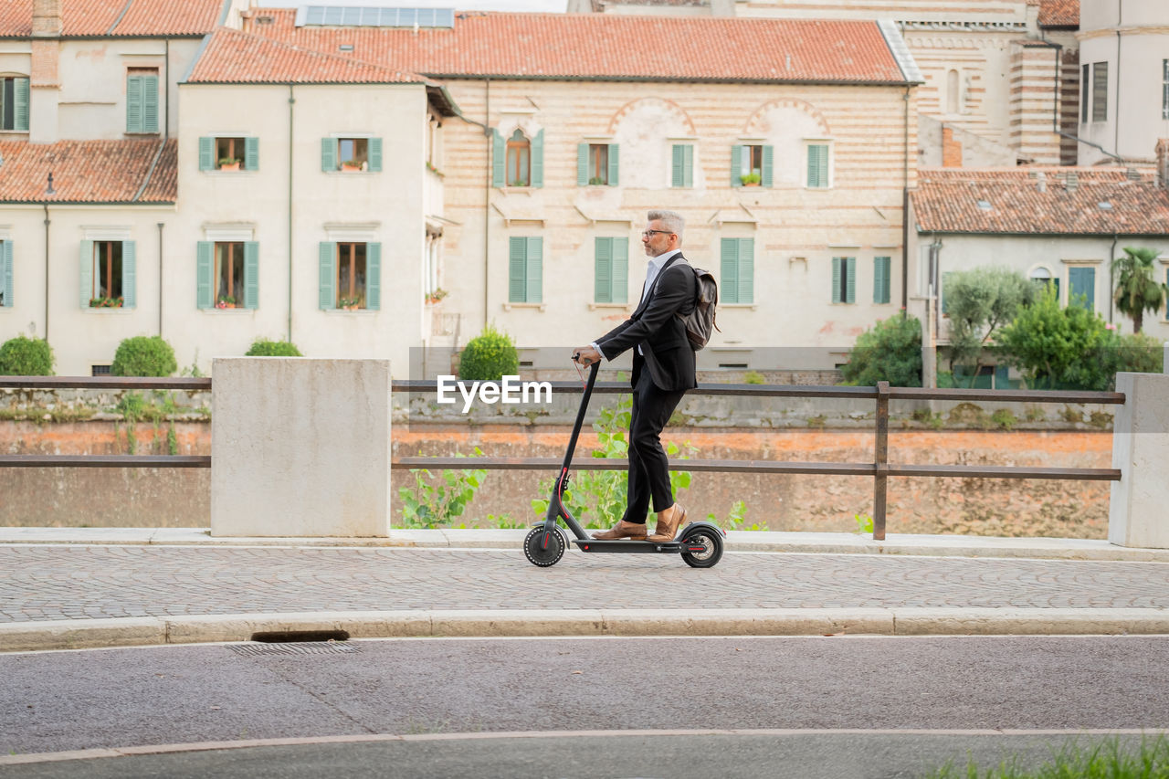 Side view full length young businessman in a suit riding an electric scooter while commuting to work
