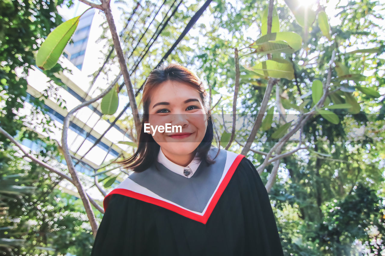 Portrait of smiling young woman wearing graduation gown against tree