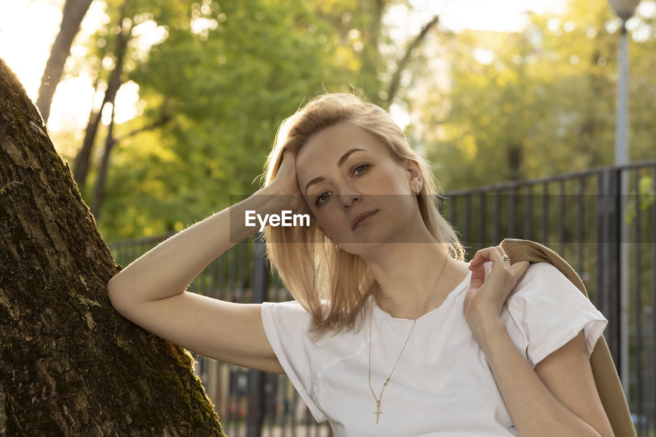 Tired caucasian real woman leans to tree, looks at camera in summer time on sunset. rest and