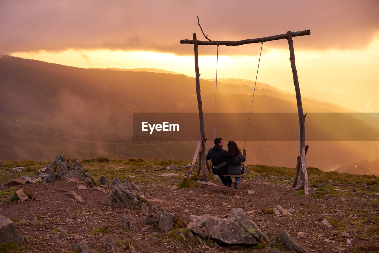 REAR VIEW OF PEOPLE STANDING ON MOUNTAIN AGAINST SKY DURING SUNSET