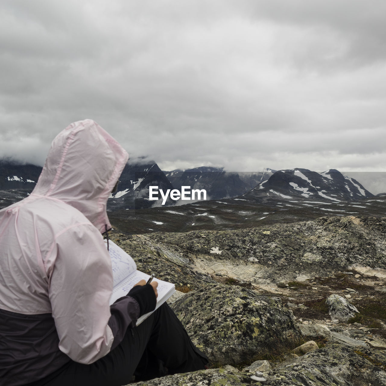 Side view of woman writing while sitting on mountain against cloudy sky