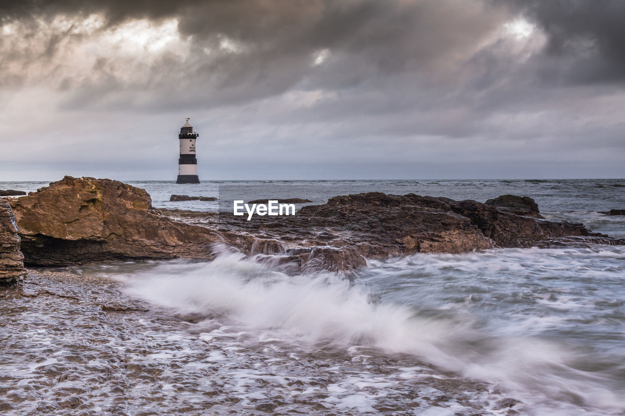 LIGHTHOUSE AMIDST ROCKS ON SEA AGAINST SKY