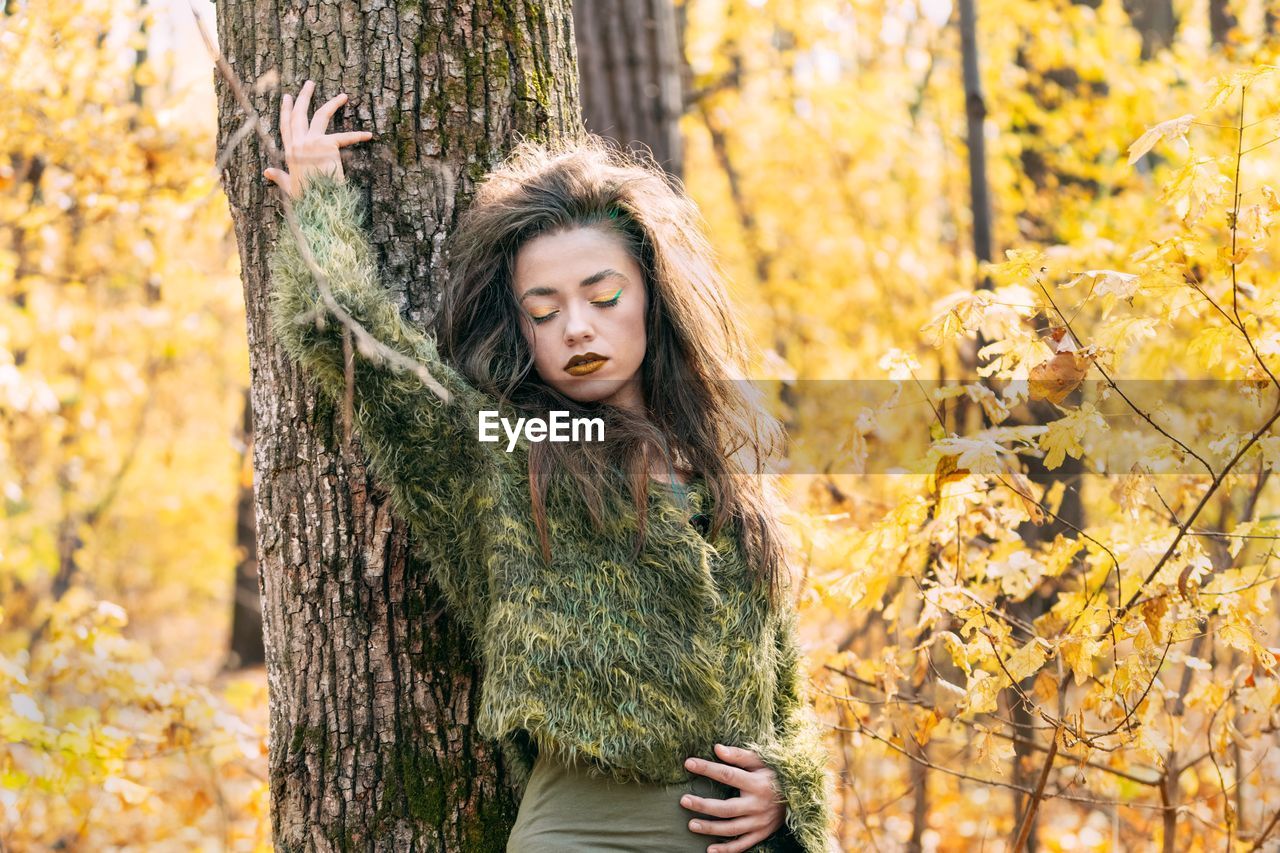 Young woman standing against tree trunk in forest during autumn