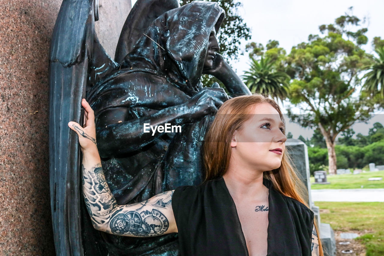 Beautiful young woman standing by statue in cemetery