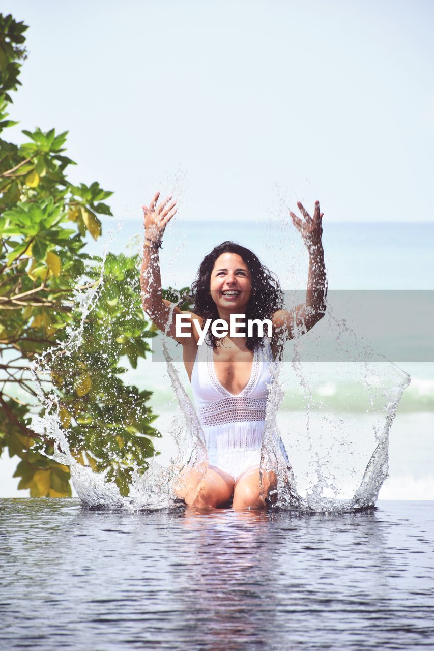 Happy young woman playing with water while sitting in infinity pool