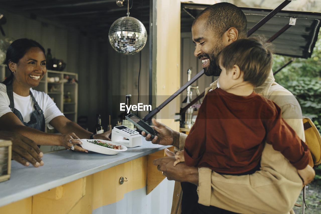 Smiling male customer with son doing contactless payment through smart phone