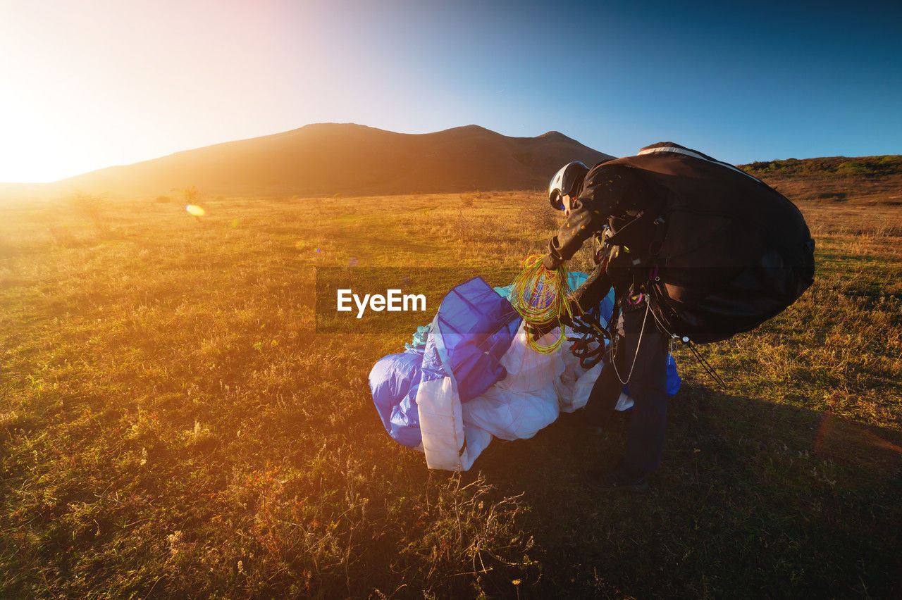 A young male paraglider at sunset in the mountains collects his paraglider to take it to the