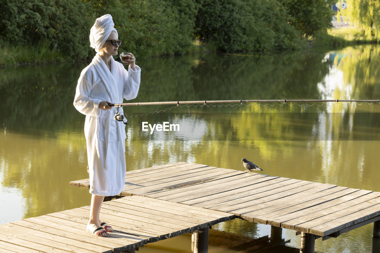 Caucasian woman in white robe and towel is fishing holding spinning rod on pier of lake early in the