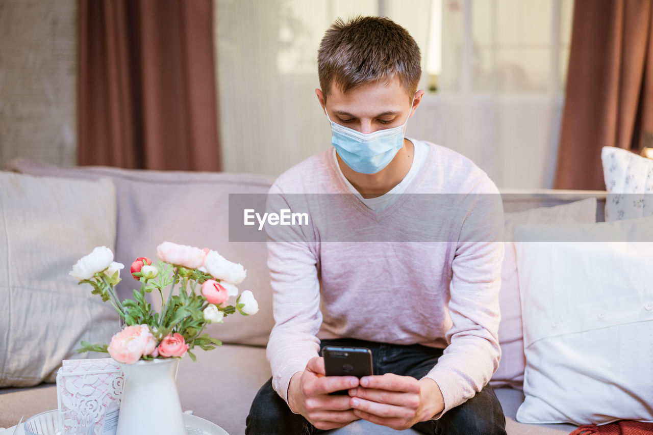 A young man sits on a sofa wearing a protective mask with a phone in his hand