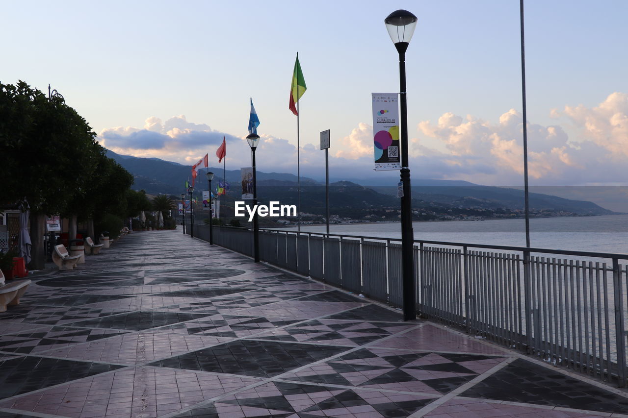 walkway, sky, water, nature, boardwalk, architecture, flag, street light, travel destinations, sea, mountain, cloud, street, city, scenics - nature, lighting equipment, no people, beauty in nature, built structure, sunset, travel, tranquility, outdoors, tourism, vacation, railing, coast, pier, patriotism, footpath, environment, tranquil scene, building exterior, day, transportation, beach