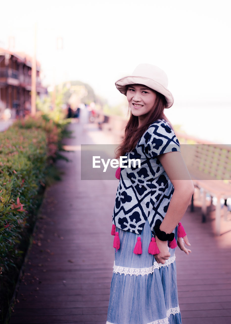 PORTRAIT OF SMILING YOUNG WOMAN IN HAT STANDING AGAINST CITY