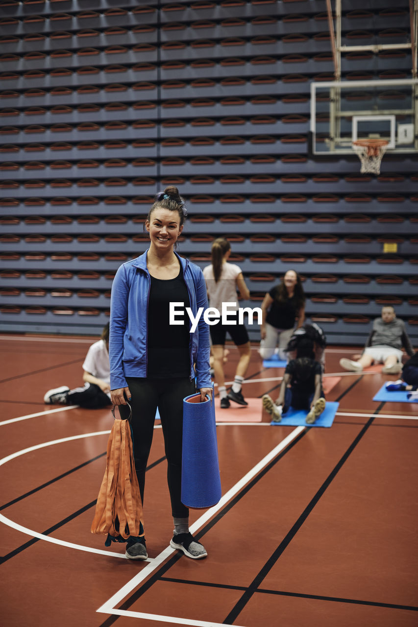 Full length portrait of smiling female coach standing with exercise mat in sports court