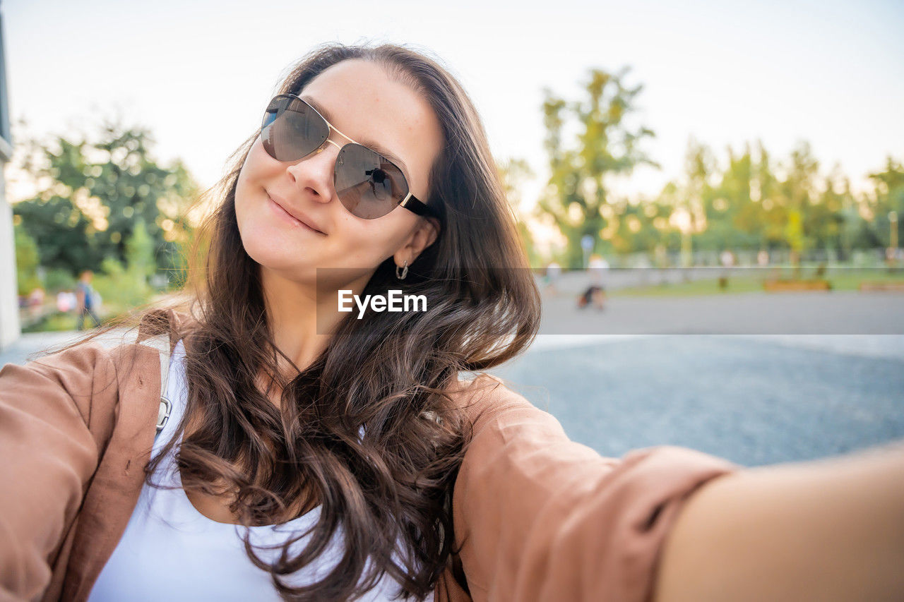 portrait of smiling young woman wearing sunglasses while sitting against trees