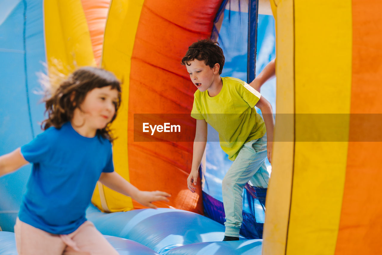 Adorable content little sister and brother with dark hair in t shirts running inside of colorful inflatable trampoline while spending time together on playground on sunny day