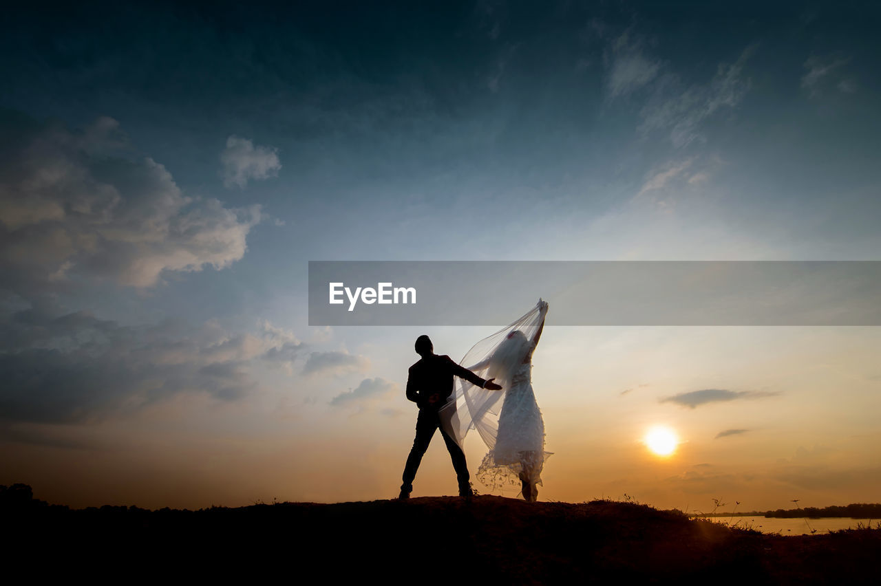 Rear view of bride and groom standing on field against sky during sunset