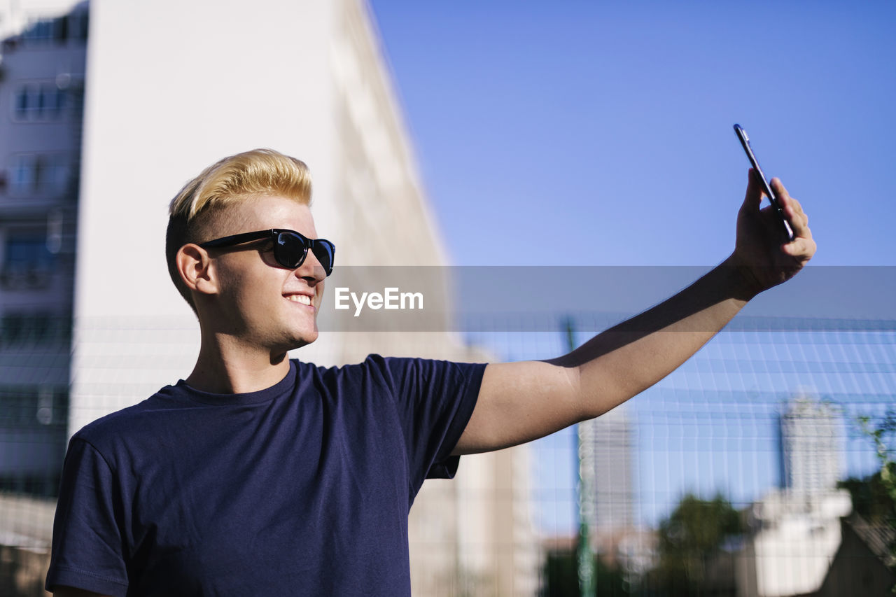 YOUNG MAN WEARING SUNGLASSES STANDING AGAINST CITY