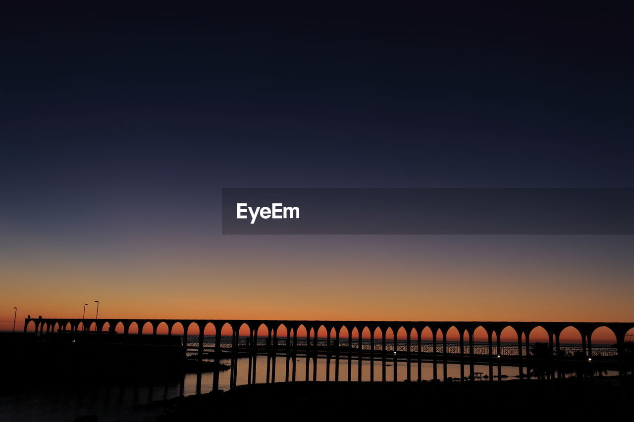 Silhouette bridge over river against clear sky during sunset