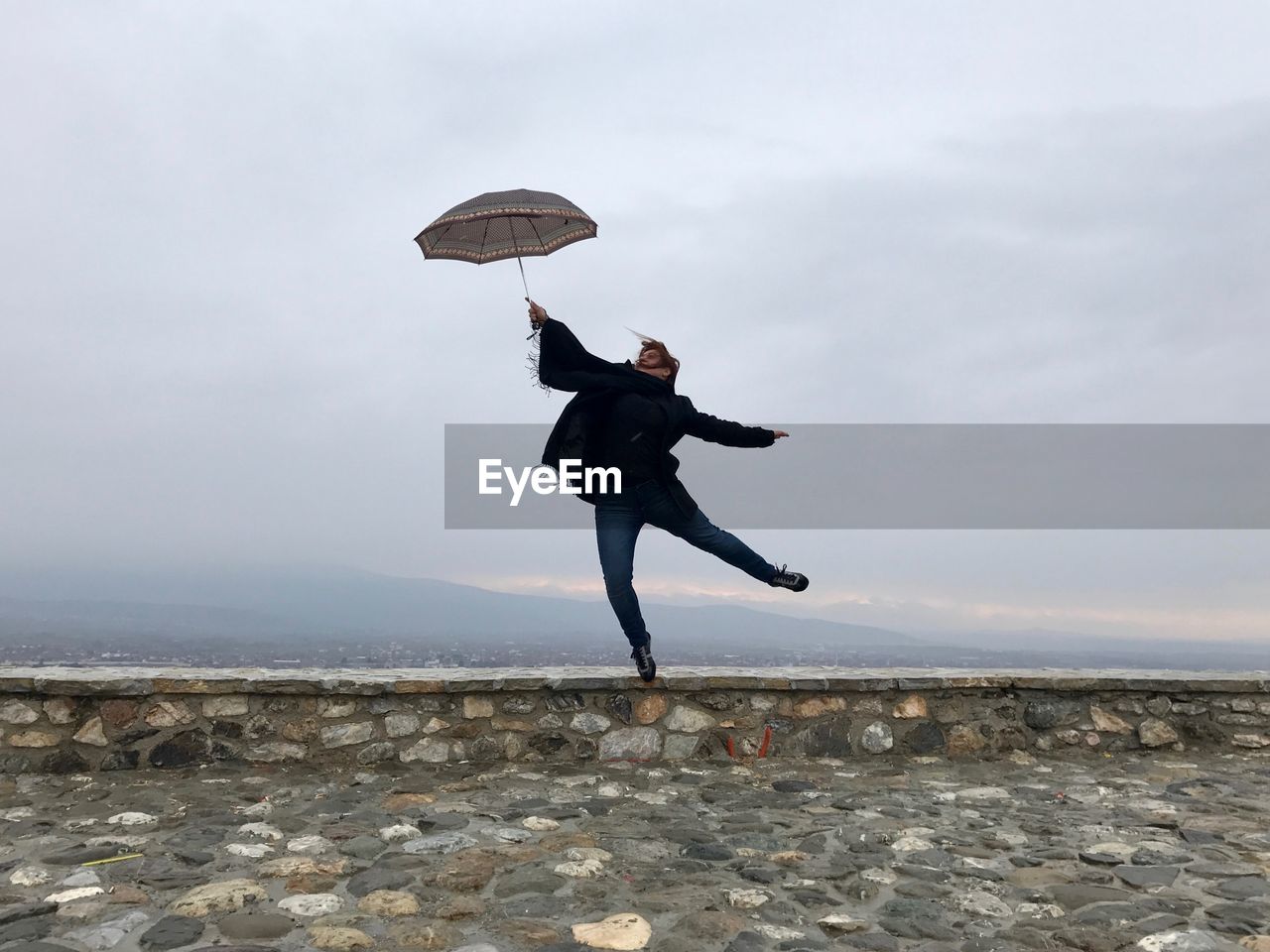 Full length of young woman jumping with umbrella on retaining wall against sky