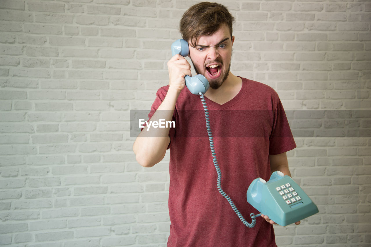 Young man yawning while using telephone against brick wall