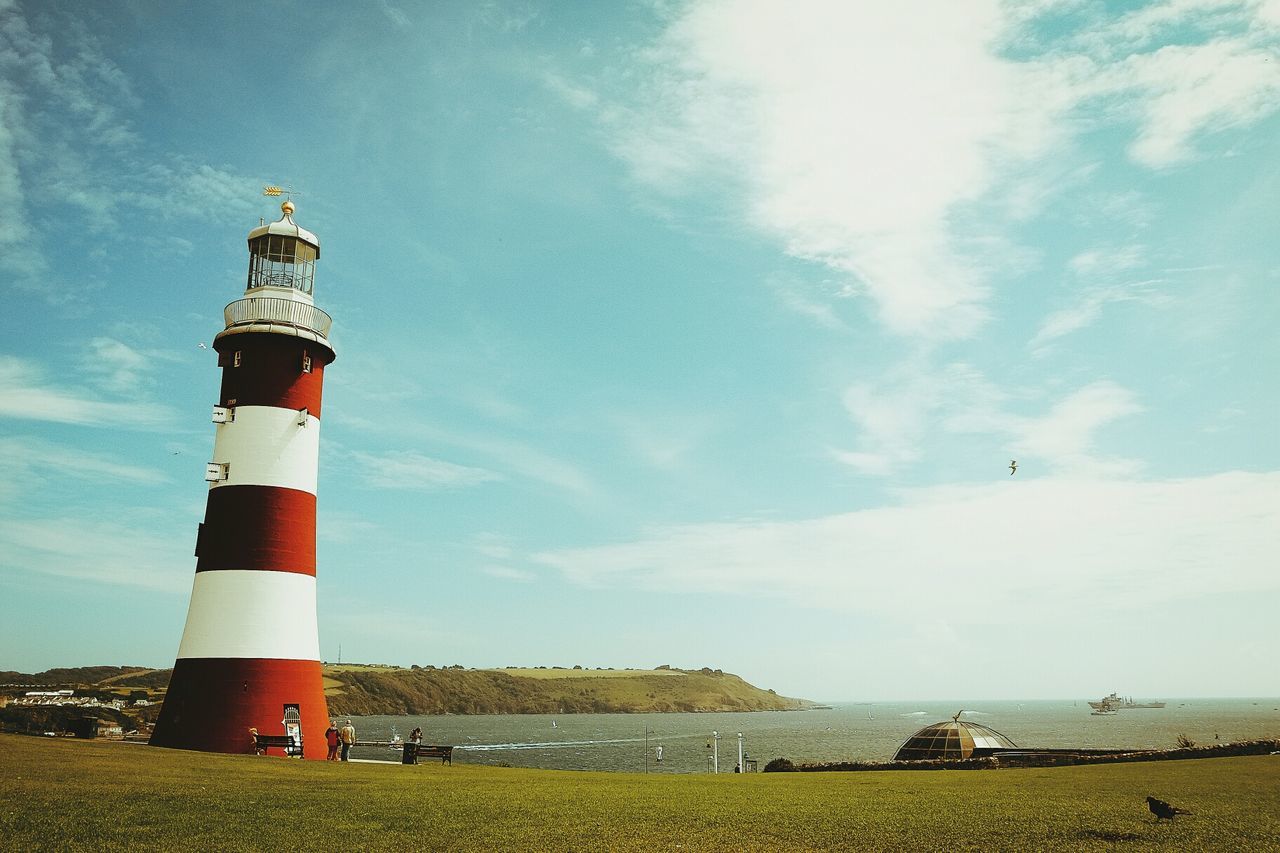 People by lighthouse on landscape against calm sea