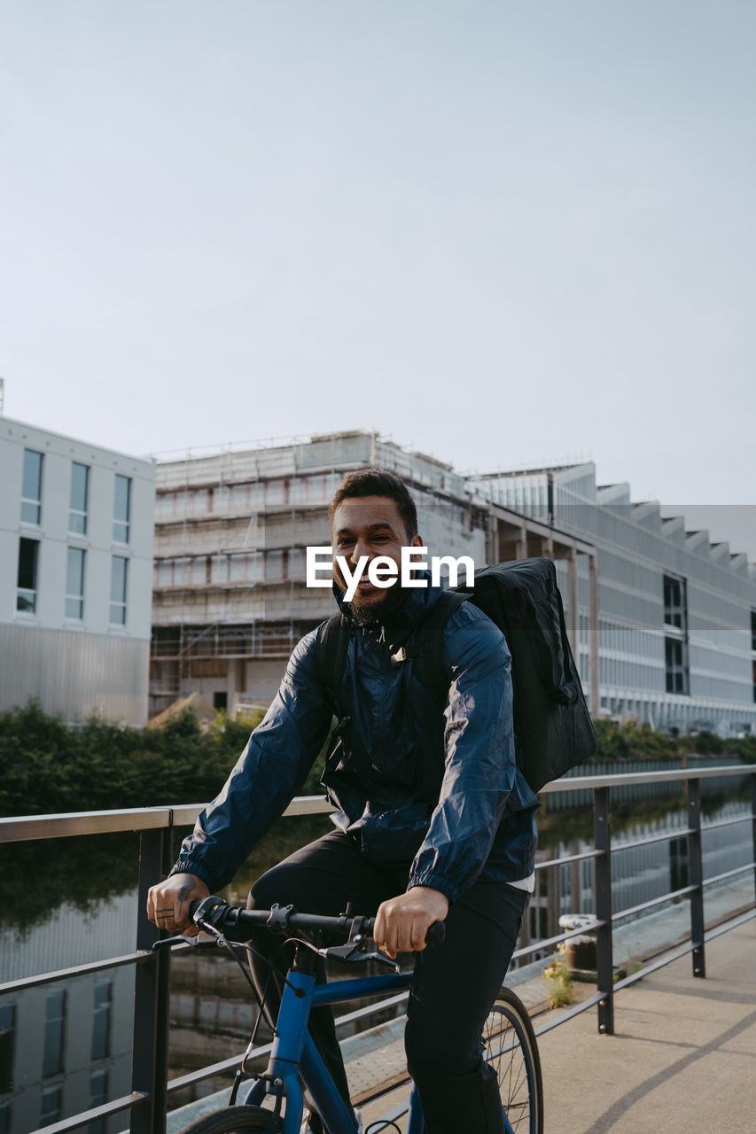 Portrait of smiling young male delivery person cycling on footpath in city