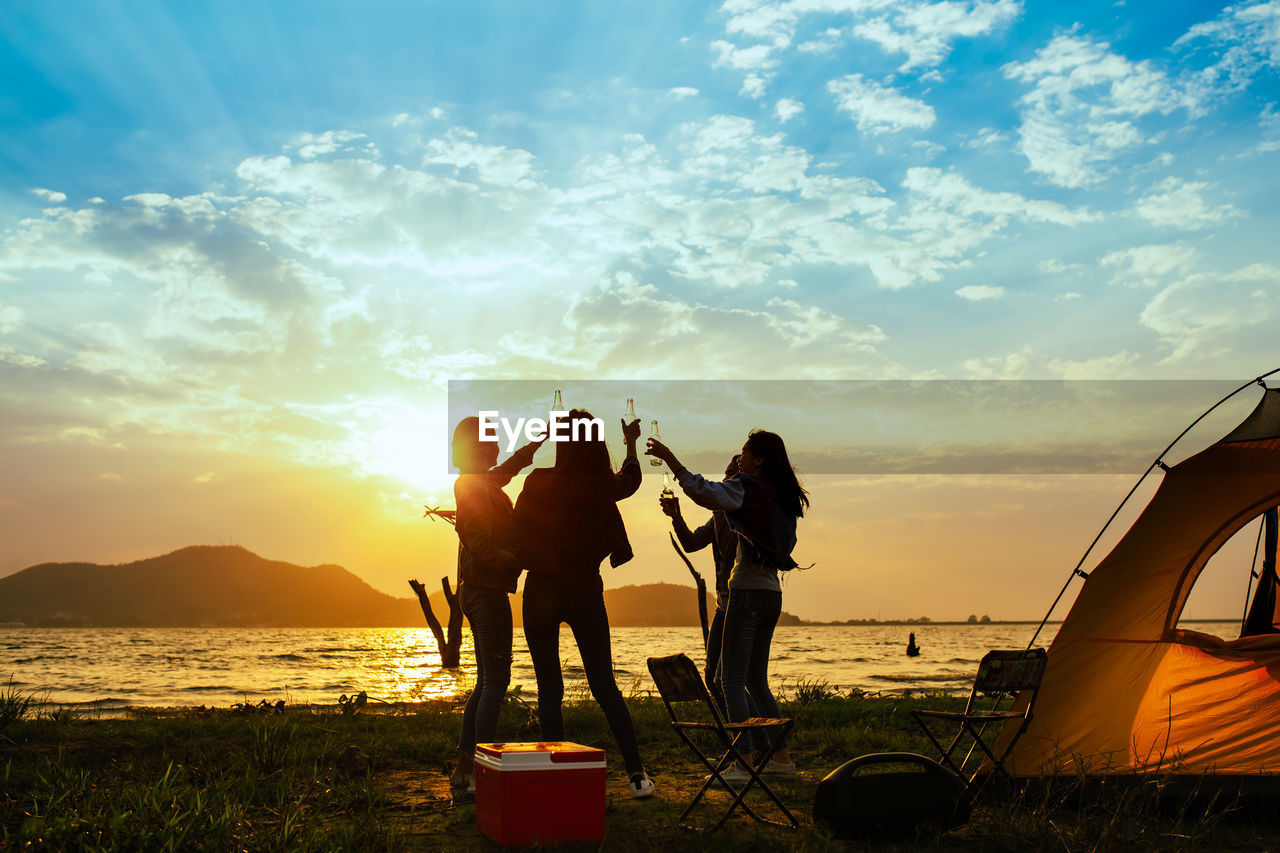 Friends partying while camping at beach against sky during sunset