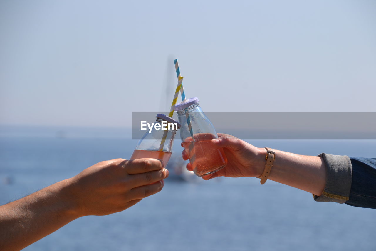 Cropped image of friends hands toasting drinks by sea against sky