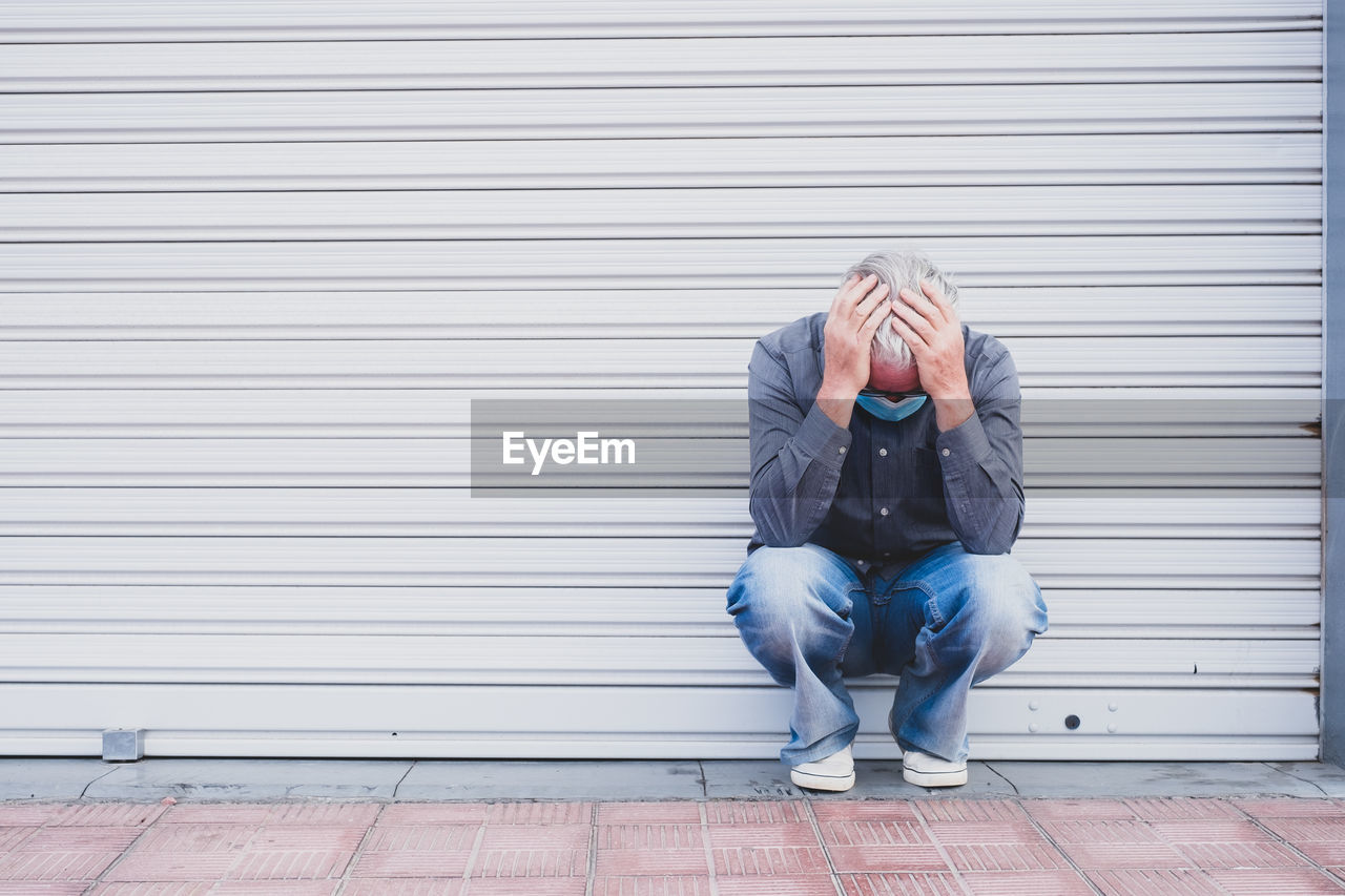 Full length of senior man in mask crouching by closed shutter