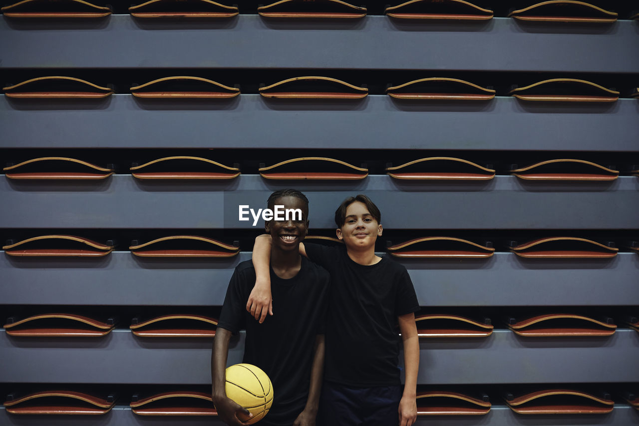 Happy teenage boy with basketball standing by male friend at sports court