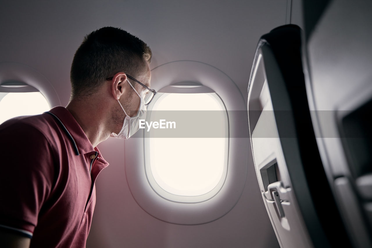 Passenger with protective face mask in airplane looking through window. 