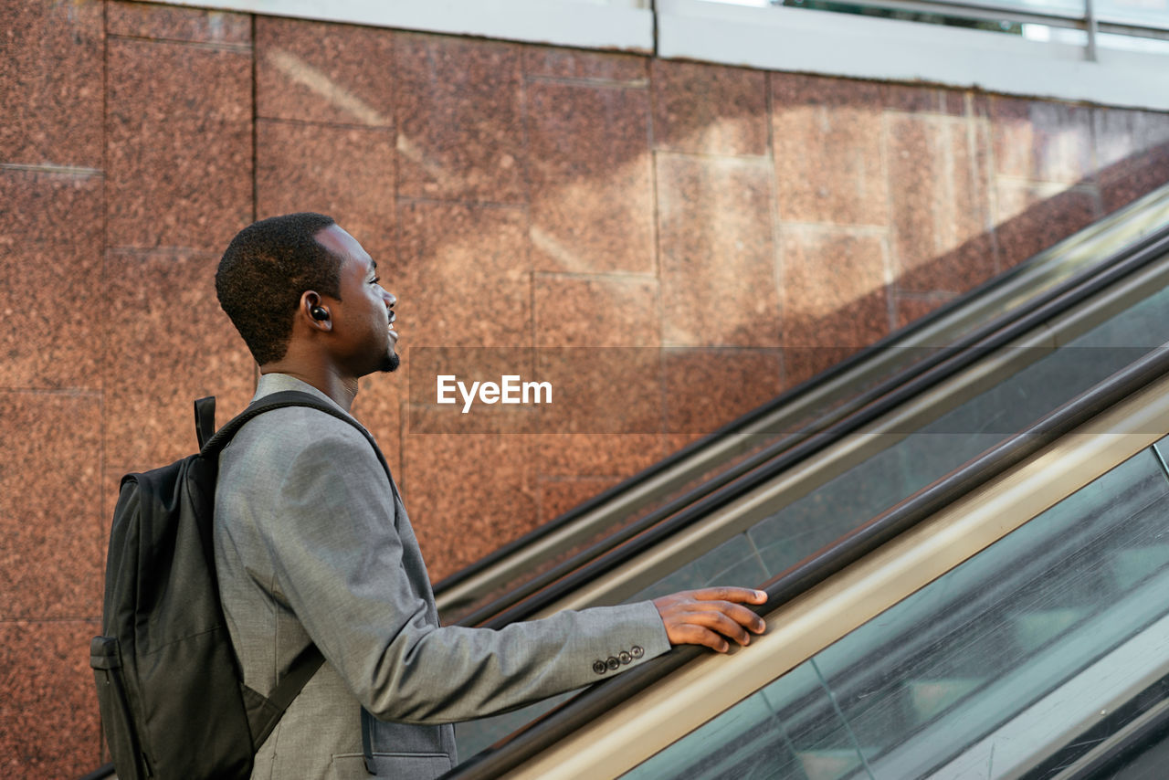 Side view of african american male entrepreneur with backpack listening to songs in tws earbuds on escalator